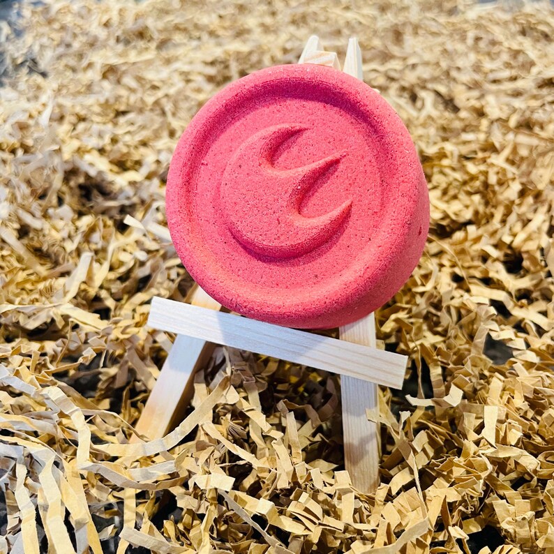 May include: A round, pink bath bomb with a stylized design, displayed on a miniature wooden easel. The bath bomb is the focal point, set against a backdrop of shredded brown paper.
