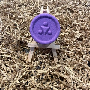 May include: A round, purple object with a textured surface and a symbol of a person above two circles and a triangle, displayed on a small wooden easel. The background is shredded brown paper.