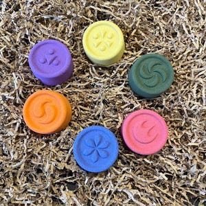 May include: Six colorful, round bath bombs with embossed designs are arranged on a bed of brown paper. The bath bombs are purple, yellow, green, orange, blue, and pink. Each bath bomb has a unique symbol design.