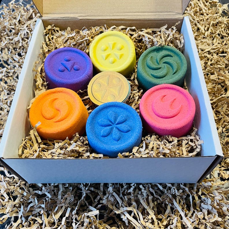 May include: A box filled with colorful, round bath bombs. The bath bombs are in shades of purple, yellow, green, orange, blue, pink, and gold. Each bath bomb has a unique embossed design, including a flower, a swirl, and a smiley face.