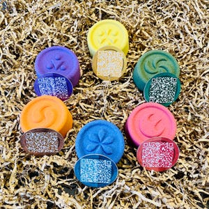 May include: A collection of colorful, round bath bombs in shades of yellow, purple, green, orange, blue, and pink. Each bath bomb has a unique embossed design and is paired with a small, round tag featuring a QR code. The bath bombs are arranged on a bed of shredded paper.