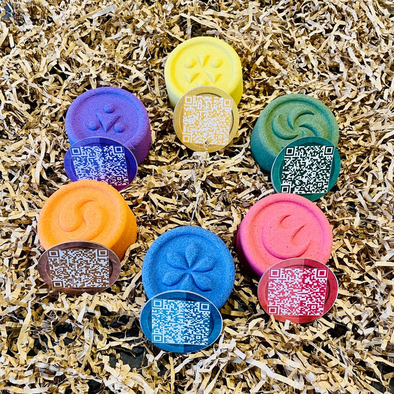 May include: A collection of colorful, round bath bombs in shades of yellow, purple, green, orange, blue, and pink. Each bath bomb has a unique embossed design and is paired with a small, round tag featuring a QR code. The bath bombs are arranged on a bed of shredded paper.