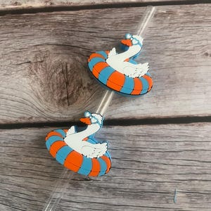 May include: Two clear straws topped with a white swan wearing sunglasses and floating on a blue and orange inner tube. The swan has an orange beak and is surrounded by a blue and orange striped floatie. The background is a light brown wooden surface.