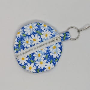 Op de afbeelding: Een ronde muntenbeurs met een madeliefjesbloemmotief op een blauwe achtergrond. De beurs heeft een witte rits en een zilveren sleutelring. Het madeliefjespatroon heeft witte bloemblaadjes en gele centra.