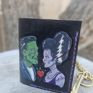 May include: Black leather key holder featuring a hand-painted image of Frankenstein and his bride, with a red heart between them. The key holder has purple stitching and a gold-colored metal key ring and clasp.