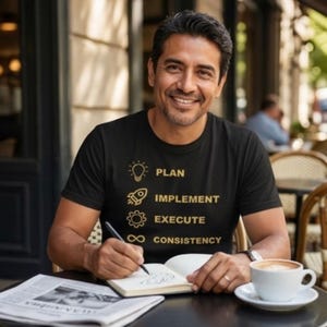 K&ouml;nnte beinhalten: Ein schwarzes Kurzarm-T-Shirt mit goldfarbenen Symbolen und Text. Das Design zeigt ein Gl&uuml;hbirnensymbol mit dem Wort PLAN, eine Rakete mit IMPLEMENT, ein Zahnrad mit EXECUTE und ein Unendlichkeitssymbol mit dem Wort CONSISTENCY.