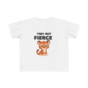 May include: A white short-sleeved t-shirt featuring a graphic of a small orange tiger with brown stripes. Above the tiger, the text reads "TINY BUT FIERCE" in bold, dark grey, rounded capital letters. The design is centred on the chest area.