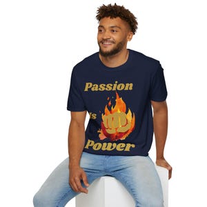 Op de afbeelding: Een marineblauw t-shirt met korte mouwen en een grafische print van een gouden vuist omringd door oranje en rode vlammen. De tekst "Passion is Power" is in gouden letters boven en onder het centrale vlam- en vuistontwerp gedrukt.