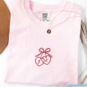 May include: A light pink t-shirt featuring an embroidered design of two red hearts tied together with a red bow. The left heart contains the letter J and the right heart contains the letter E. The shirt has a Comfort Colors brand label at the neck.