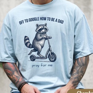 May include: Light blue t-shirt with a raccoon graphic riding a scooter. The text "OFF TO GOOGLE HOW TO BE A DAD" is above the raccoon, and "pray for me" is below. The shirt is labeled "Chambray".