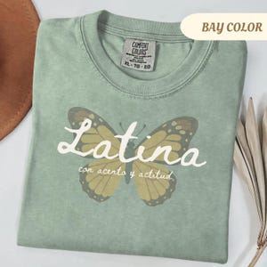 May include: A sage green t-shirt with a butterfly graphic and the word "Latina" in white script. The butterfly has brown and yellow details. The text "con acento y actitud" is below the word "Latina".