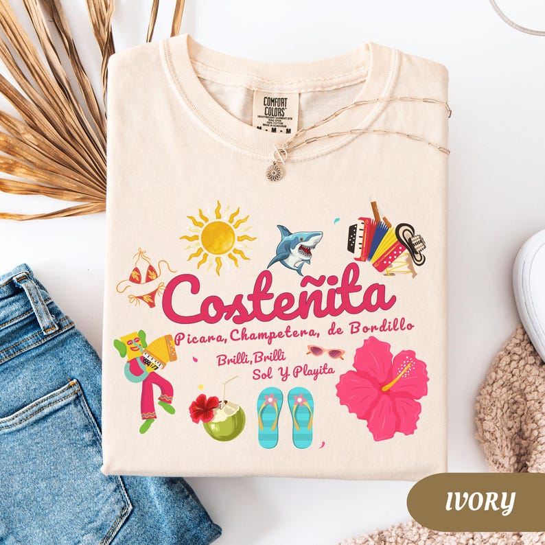 May include: Ivory-colored t-shirt with a graphic design featuring the word "Coste&ntilde;ita" in pink, along with illustrations of a sun, shark, bikini, accordion, and hibiscus flower. The shirt also has the text "Picara, Champetera, de Bordillo, Brilli, Brilli, Sol Y Playita."