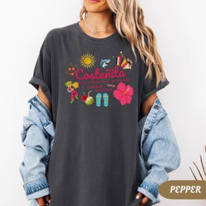 May include: Dark gray t-shirt with a colorful graphic design. The design includes the word "Coste&ntilde;ita" in pink, along with illustrations of a sun, shark, musical instruments, flowers, and other tropical elements. The shirt is a relaxed fit.