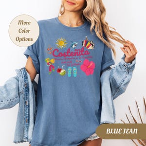 May include: Blue jean colored t-shirt with a colorful graphic design. The design includes the word "Coste&ntilde;ita" with illustrations of a sun, shark, musical instruments, flowers, and other tropical elements. The shirt has rolled-up sleeves.