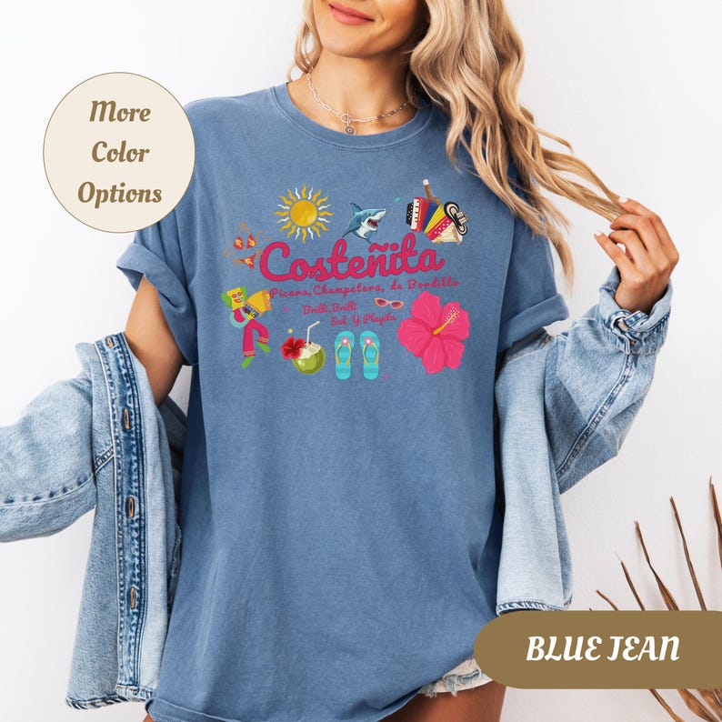 May include: Blue jean colored t-shirt with a colorful graphic design. The design includes the word "Coste&ntilde;ita" with illustrations of a sun, shark, musical instruments, flowers, and other tropical elements. The shirt has rolled-up sleeves.
