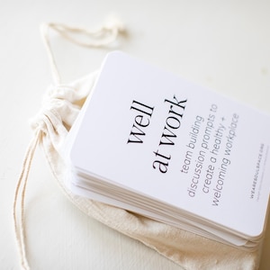 May include: A stack of white cards with rounded corners, printed with the words "well at work" and other text, are inside a beige drawstring bag. The cards are designed to help build teams and create a welcoming workplace.