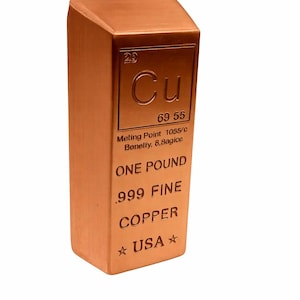 May include: A copper bar with the element symbol Cu, atomic number 29, and the text "ONE POUND .999 FINE COPPER USA". The bar has a rectangular shape with a slanted top and is a reddish-brown color.