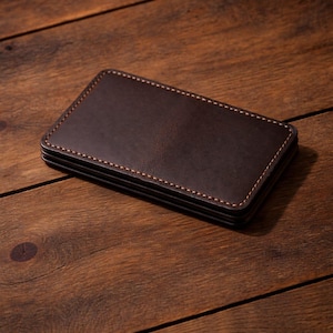 Può includere: Un set di quattro sottobicchieri in pelle marrone scuro con cuciture marrone chiaro. I sottobicchieri rettangolari sono appoggiati su una superficie di legno.