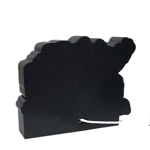 Puede incluir: Una luz decorativa negra con forma de nube con una superficie similar al fieltro. La luz tiene una base rectangular y un cable blanco que se extiende desde la parte inferior. La parte superior de la luz tiene un dise&ntilde;o ondulado en forma de nube.