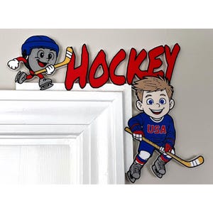 Puede incluir: Un letrero decorativo de madera con la palabra "HOCKEY" en rojo, y jugadores de hockey de dibujos animados. Uno es un disco gris con casco, el otro es un niño con una camiseta azul "USA". El letrero está diseñado para un marco de puerta.