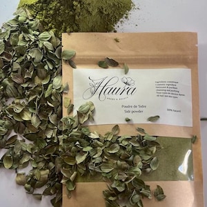 May include: A brown paper bag labeled "Haura" containing green powder and dried leaves. The bag is partially transparent, showing the green powder inside. A pile of green powder and dried leaves are scattered around the bag.