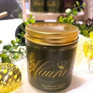 Peut inclure: Un pot en verre transparent avec un couvercle doré, rempli de poudre vert foncé. Le pot porte une étiquette vert foncé avec l'inscription dorée "Haura Herbs & Beauty" et "Poudre de Katam Katam powder".