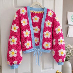 May include: A bright pink crochet cardigan with white and yellow daisy flower appliques. The cardigan has a light blue trim and a tie closure at the waist. The sleeves are long and puffy.