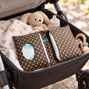 May include: A brown fabric pouch with white polka dots designed for baby wipes, featuring a central oval opening. It is placed inside a stroller alongside a beige plush rabbit toy, a baby bottle with a tan cap, a muslin cloth with bear prints, and a wooden ring toy.