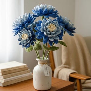 Puede incluir: Un ramo de flores azules hechas a mano con ganchillo en un jarrón de cerámica blanco. Las flores tienen pétalos de diferentes tonos de azul y un centro amarillo claro. El jarrón está sobre una mesa de madera.