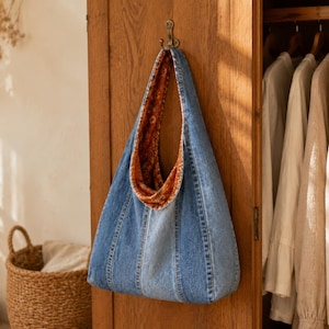 Puede incluir: Un bolso de hombro estilo hobo confeccionado con paneles de tela vaquera azul. El forro interior presenta un tejido con un estampado vibrante en tonos naranja y marrón. El bolso cuelga de un gancho metálico, mostrando su silueta holgada.