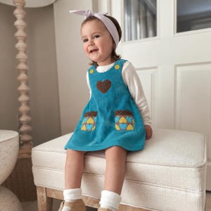 May include: A teal corduroy pinafore dress featuring a brown heart appliqué with white polka dots on the chest, two front pockets with a yellow and blue heart pattern, and yellow buttons at the shoulders, worn over a long-sleeved white shirt.