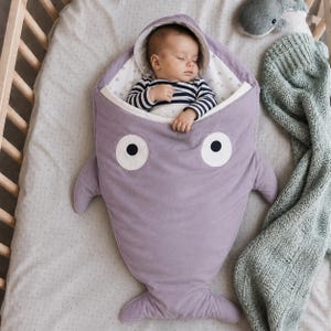 Peut inclure: Une gigoteuse pour bébé lavande en forme de baleine avec de grands yeux circulaires blancs et noirs. La gigoteuse présente une capuche et des nageoires latérales. Un bébé portant un haut à manches longues rayé bleu marine et blanc est installé.