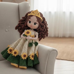 Pode incluir: Boneca de crochê feita à mão com cabelo castanho encaracolado, vestindo um vestido creme adornado com girassóis amarelos e uma bainha verde. A boneca tem uma coroa amarela e segura um girassol. A boneca está sentada em uma poltrona cinza.