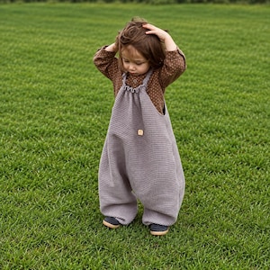 May include: A waffle-textured, light purple overall with a gathered neckline and a small square tag on the chest, worn over a brown long-sleeved shirt with white polka dots and dark blue shoes, standing on a green grass field.
