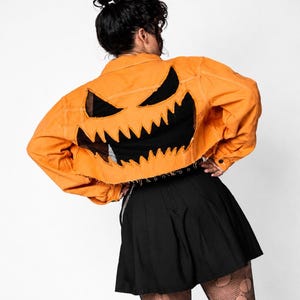Può includere: Una giacca di jeans corta color arancione con un grande inserto in tessuto nero dai bordi frastagliati sul retro, che forma un volto di zucca stilizzato con occhi triangolari appuntiti e un ampio sorriso dentato. La giacca presenta un orlo sfrangiato.