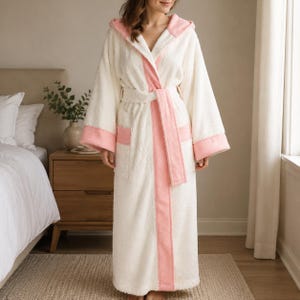 Può includere: Un accappatoio lungo in spugna bianca testurizzata con cappuccio, maniche ampie e cintura in vita coordinata. L'accappatoio è rifinito con un bordo rosa tenue lungo l'apertura frontale, i polsini, il cappuccio e le due tasche anteriori.