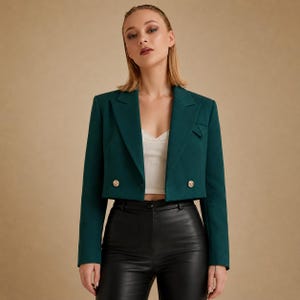 May include: A cropped dark green blazer featuring long sleeves, a notched lapel, and two gold-toned buttons on the front. The blazer is worn over a white ribbed tank top and black trousers, showcasing a structured design with clean lines.
