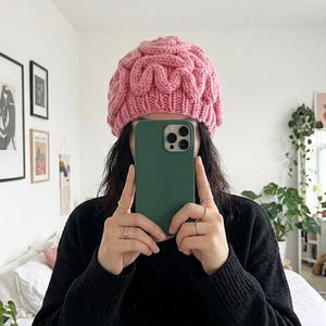 May include: A chunky knit beanie in a soft pink color featuring a raised, swirling brain-like texture pattern throughout the crown. The hat has a ribbed brim and is worn on the head, paired with a black long-sleeved sweater and a dark green smartphone.