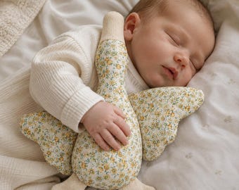 May include: A plush toy shaped like a goose with a floral pattern featuring small yellow flowers and green leaves on a white background. The toy has solid cream-colored fabric on the head and feet, and it is held by a sleeping infant in a cream shirt.
