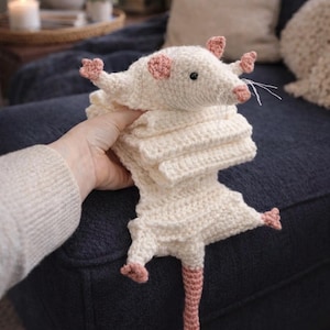 May include: A handmade crochet rat plushie in cream with pink accents. The rat has a long, pink tail, pink ears, and tiny pink feet. The plushie is lying on a dark blue couch.