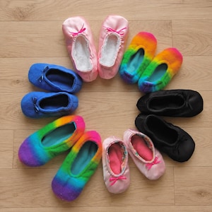 Peut inclure: Une collection de dix chaussons souples disposés en cercle, présentant des couleurs et des textures variées. L'ensemble comprend des paires en satin rose uni, en polaire bleue unie, en polaire arc-en-ciel, en tissu noir et en satin rose clair.
