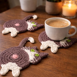 May include: Crocheted cat-shaped coasters in shades of brown, white, and pink. The coasters have green button eyes and are arranged on a wooden surface. A white coffee cup sits on one of the coasters.