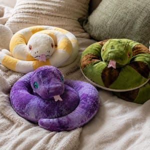 May include: Three plush snake toys coiled in circular shapes. One snake is white and yellow, one is purple with a textured finish, and one is green and brown with a rattle on its tail. Each snake features a small pink tongue and round eyes.
