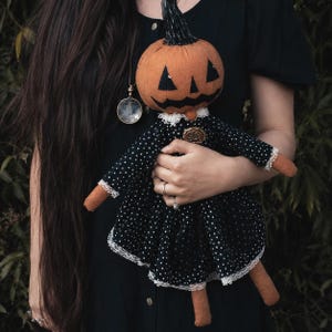 Może przedstawiać: Szmaciana lalka z pomarańczową głową dyni, czarną malowaną twarzą jack-o'-lantern i czarną łodygą. Lalka ma na sobie czarną sukienkę w białe kropki, z białym koronkowym wykończeniem przy kołnierzu i mankietach oraz małą złotą broszką.