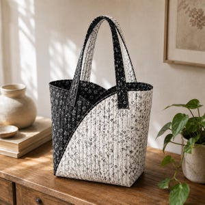May include: A quilted tote bag featuring a diagonal color-blocked design with a black section on the left and a white section on the right, both adorned with a small black floral pattern. The bag has two matching quilted handles and vertical stitching.