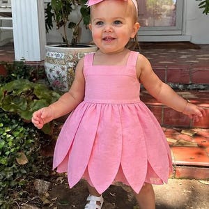 May include: A light pink sleeveless dress featuring a square neckline and a skirt constructed from overlapping petal-shaped fabric panels. The outfit includes a matching large pink hair bow attached to a thin headband and white strappy sandals.