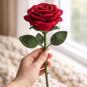 May include: A handcrafted, red crochet rose with green leaves and stem. The rose is held up, showcasing the detailed stitching and realistic design. This unique, handmade flower is a charming decorative item.