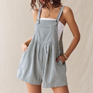 May include: A grey and white vertical striped playsuit featuring a square neckline, wide shoulder straps with button fastenings, a front chest pocket, and two side pockets. The garment is worn over a plain white tube top, creating a layered look.