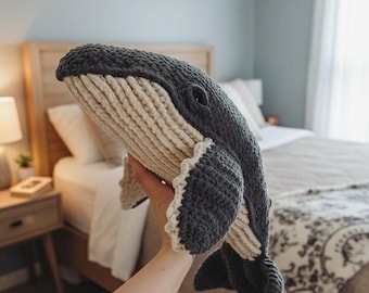 May include: A handmade, crocheted whale plushie in shades of gray and cream. The whale features detailed stitching, a textured body, and a curved tail. The plushie is held up in front of a bed.