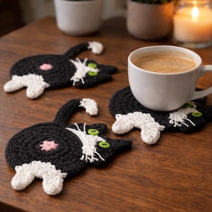 May include: Handmade crochet cat-shaped coasters in black and white with green eyes and pink accents. One coaster holds a white coffee cup. The coasters are on a wooden surface.
